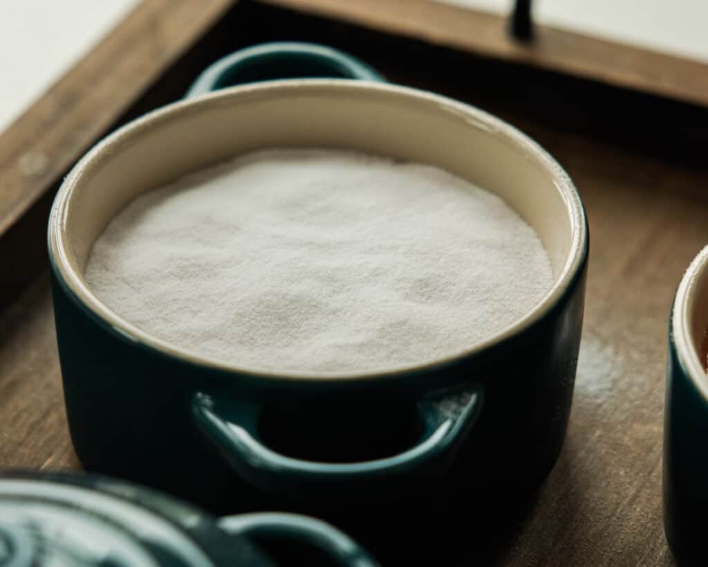 A round teal ceramic dish filled with a fine white powder, likely baking soda or sugar, sits on a wooden tray—perfect for whipping up easy vegan treats like Pink Strawberry Crème Brûlée.